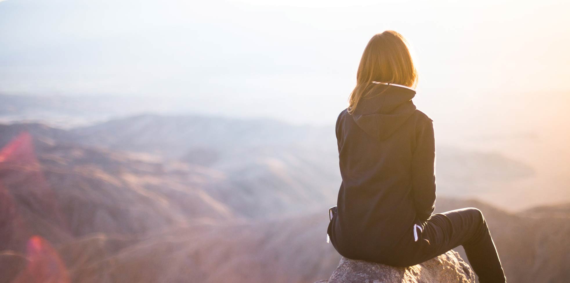 Eine Frau sietzt auf einem Stein und blickt nachdenklich in den Sonnenuntergang